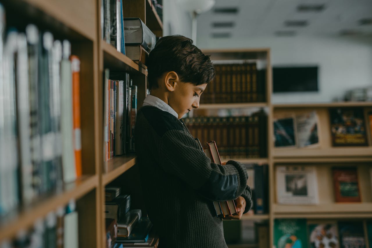 A boy looking sad and hugging books while leaning on a bookcase in a library